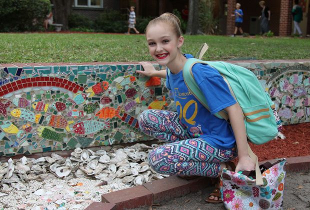Fifth-grader Burlin Frohock poses next to the school of fish created by her class for the mosaic mural on the front lawn of Fishweir Elementary School.
