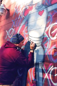 Mural artist Christopher Sweeney puts finishing touches on a colorful mural of his signature stretched hearts and popular XoRobot on the side of the CareMax Pharmacy at the corner of Park and James Streets.