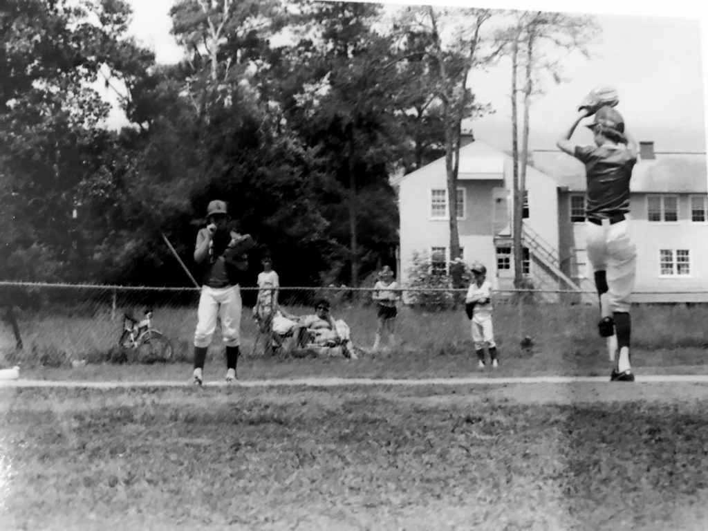 Parents sit in the weeds behind a chain-link fence to watch their sons play Little League baseball in the 1970s.
