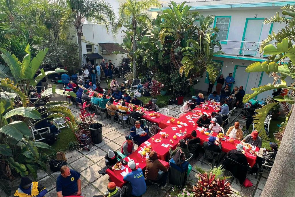 people eating outside a building at red-covered, criss-cross tables with palm trees by the building