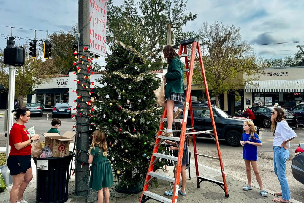 2 women and children around a Christmas tree outside, with a girl on a ladder adding a decoration