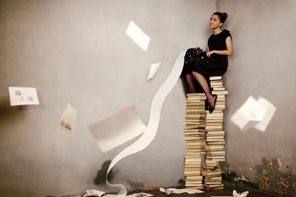 Woman on a tall stack of books, typing on a typewriter with long, flowing paper rolling down from it, and papers fluttering in the air and falling to the ground.