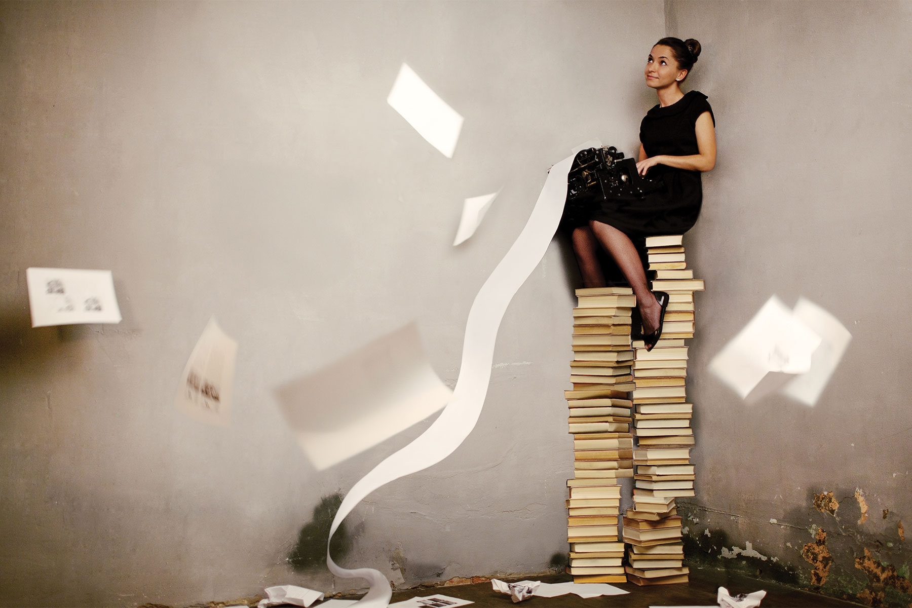 Woman on a tall stack of books, typing on a typewriter with long, flowing paper rolling down from it, and papers fluttering in the air and falling to the ground.